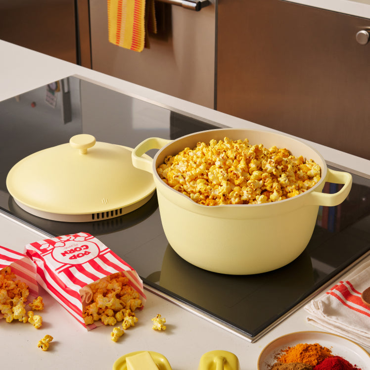 Yellow popcorn popper filled with popcorn on a stovetop with a red and white striped bag of popcorn next to it.