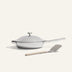 The Titanium Always Pan Pro, a white frying pan with a metal handle and advanced nonstick technology, sits beside a beige-handled spatula on a light background.