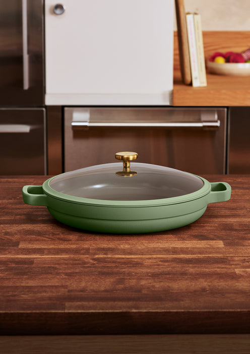 A green nonstick pan with a glass lid and gold handle sits on a wooden kitchen countertop.
