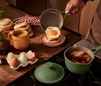 A person places a fried egg on an English muffin sandwich at a kitchen counter with tea, eggs, and a pot of simmering ingredients on the stove.