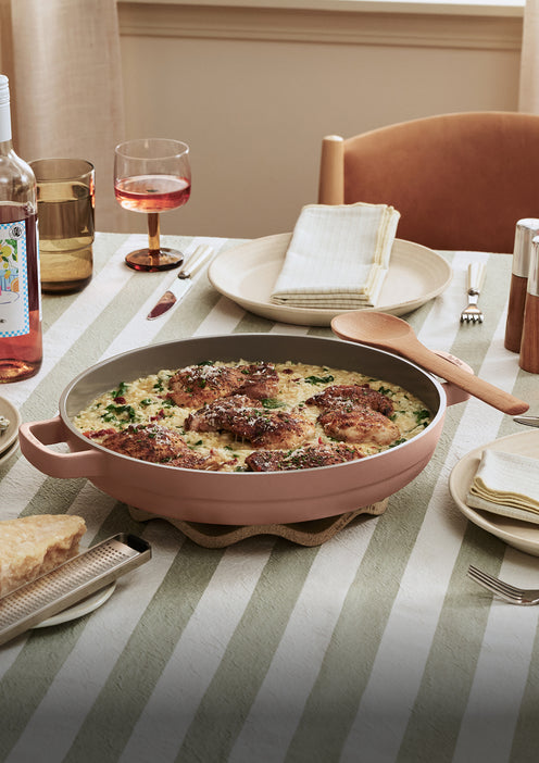 A pan of seasoned chicken and rice sits on a striped tablecloth with plates, cutlery, a wine glass, a napkin, and a bottle of rosé wine arranged nearby.