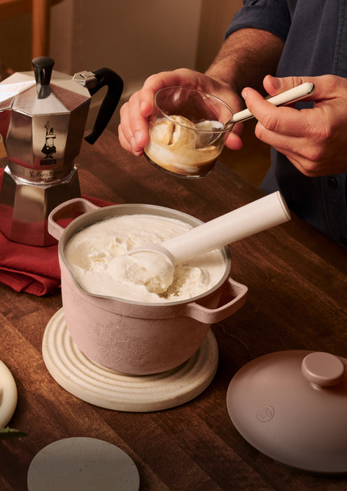 A person scoops vanilla ice cream from a pot into a glass cup, with a moka pot and a red napkin on the wooden table.