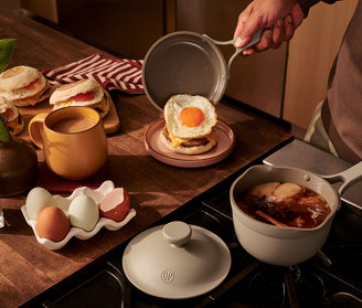 Person preparing a breakfast dish with an egg on top of a sandwich, surrounded by coffee, eggs, and a pot on a wooden table.
