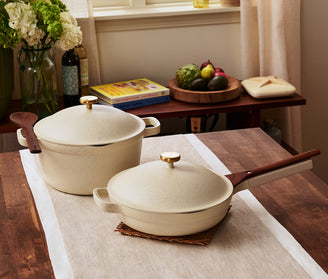 Two cream-colored ceramic cookware pieces, a pot with a lid and a pan with a lid, are placed on a wooden dining table with a light runner in a home kitchen setting.