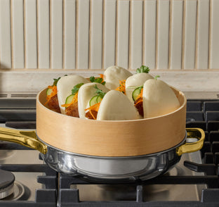 A bamboo steamer containing filled bao buns sits on top of a stainless steel pot on a stovetop.