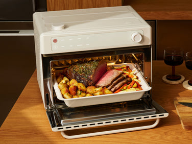 A white countertop oven with its door open, containing a baking dish filled with roasted meat, potatoes, and vegetables.