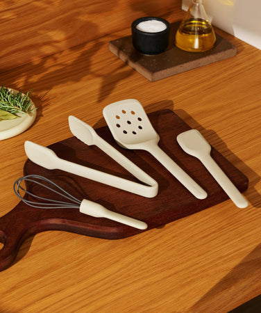A set of cream-colored kitchen utensils—tongs, a slotted turner, a spatula, and a whisk—arranged on a wooden cutting board next to a plate of herbs.
