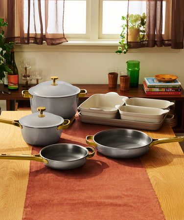 A set of gray cookware including a lidded Dutch oven, a lidded saucepan, two skillets, and a stack of ceramic baking dishes on a wooden table.