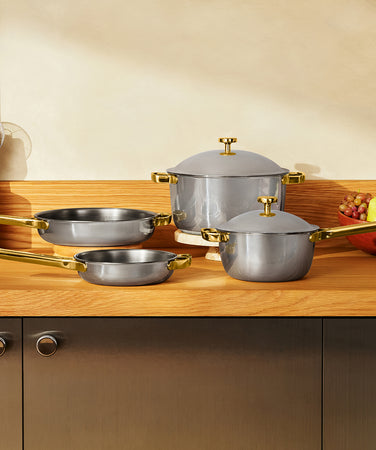 A set of gray nonstick cookware with gold-colored handles and lids arranged on a wooden table.