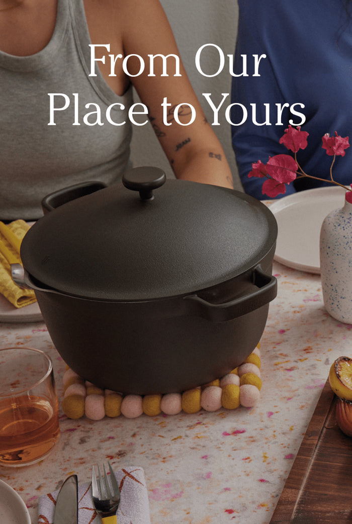Translation missing: en.A black Dutch oven sits on a table set for a meal, surrounded by plates, drinks, and a vase with pink flowers. Text reads, From Our Place to Yours