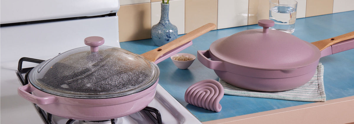 Translation missing: en.Two pink ceramic pans, one with a glass lid, sit on a blue kitchen counter next to a stove; a cup, silicone holder, and small vase are also visible