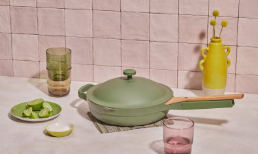 Translation missing: en.A green pan with a lid and wooden handle sits on a placemat beside a plate of lime wedges, a small bowl, glasses, and a yellow vase on a tiled surface
