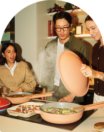 Three people stand around a kitchen island cooking with pink pans, with one person lifting a steaming lid while the others watch.