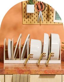 White pots, pans, and lids are organized upright in a rack on a reddish-orange countertop, with utensils hanging on a pegboard against a peach-colored wall in the background.