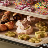 Two baking sheets filled with various cookies, including sprinkle-covered round cookies, pastries, meringues, and green-and-tan checkered squares.