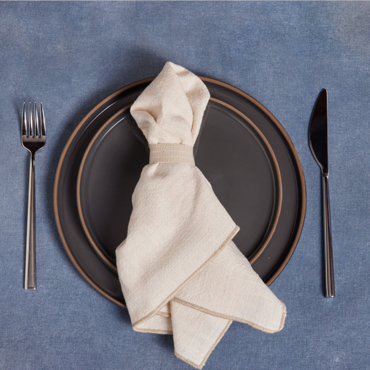 A beige Loop Napkin rests on top of black dinnerware on a blue tablecloth