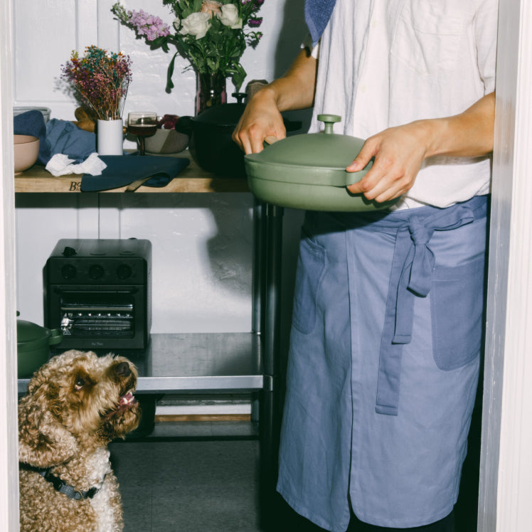 A chef wearing a blue Home Cook Apron holds a green Always Pan as a dog watches