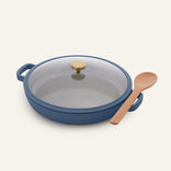 Introducing the Dual Handle Always Pan—a versatile blue nonstick pan with side handles, paired with a matching glass lid topped by a gold knob and accompanied by a wooden spoon. Crafted from non-toxic materials, this stylish pan is thoughtfully designed to bring both elegance and functionality to your kitchen.