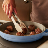A person uses tongs to cook meatballs in a light blue skillet on a stovetop.