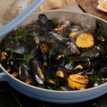 Mussels steamed in a blue pan with herbs and halved grilled lemons, releasing steam.