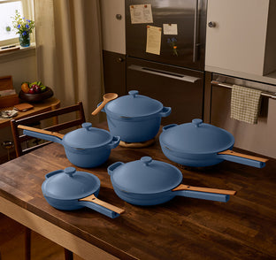 Four blue pots and pans with wooden handles and lids are arranged on a wooden kitchen table in a home kitchen setting.