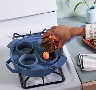 A person pours a cracked egg into a round blue silicone egg poacher insert placed on a blue pan over a gas stove.