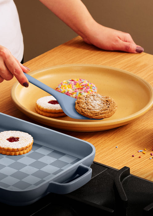 A hand uses a spatula to place a cookie onto a plate with other cookies, next to a baking pan.
