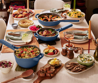 A dining table set with various dishes in blue cookware, including rice, vegetables, bread, and salads, with drinks, plates, and utensils arranged for a meal.