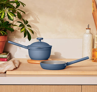 A blue saucepan with a lid and a matching blue frying pan rest on a wooden countertop beside a plant, a towel, books, and a bottle of oil.