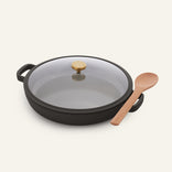The Dual Handle Always Pan, featuring a black, round nonstick design with a glass lid and gold knob, is placed next to a wooden spoon on a plain light background, showcasing its versatile design for everyday cooking.