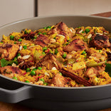 A close-up of a pan filled with chicken biryani, featuring rice, chicken pieces, green peas, spices, and garnished with sliced almonds.