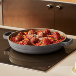 A round, gray pan filled with spaghetti, tomato sauce, and meatballs sprinkled with grated cheese sits on a kitchen stovetop.