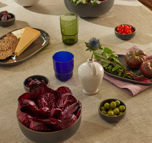 A table set with assorted foods including red lettuce, olives, cheese, crackers, tomatoes, dates, red peppers, and assorted glasses, with a small vase holding a single flower.