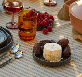 A table set with plates, spoons, glasses of red drink, a bowl of raspberries, and a dish of dates arranged around a round cheese on a striped tablecloth.