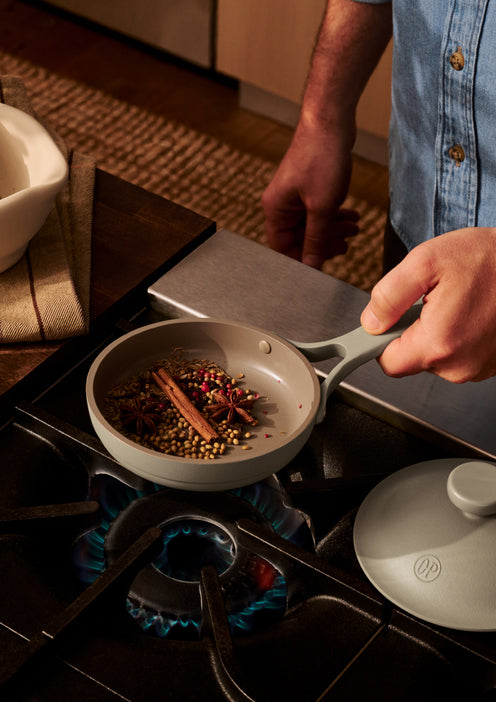 A person holds a small pan over a gas stove, toasting whole spices such as cinnamon sticks, star anise, and coriander seeds.