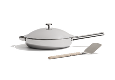 A stainless steel frying pan with a lid and a metal spatula with a beige handle placed beside it on a white background.