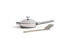 A stainless steel frying pan with a lid and a metal spatula with a light-colored handle, set against a plain white background.
