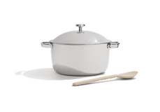 A stainless steel pot with a lid and metal handles is placed next to a beige wooden spoon on a white background.