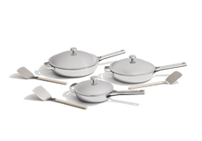 Three white cookware pans with matching lids and three light-colored spatulas are arranged on a white background.