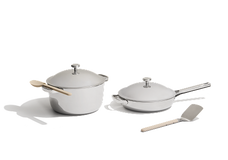 Two stainless steel cookware pieces with lids, a pot with a wooden spoon resting on it, a frying pan, and a metal spatula with a wooden handle, all on a white background.