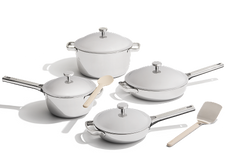 A set of four stainless steel cookware pieces with lids, a wooden spoon, and a spatula arranged on a white background.