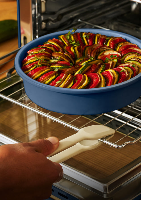 A person uses tongs to pull a blue dish containing a layered vegetable bake out of an oven.