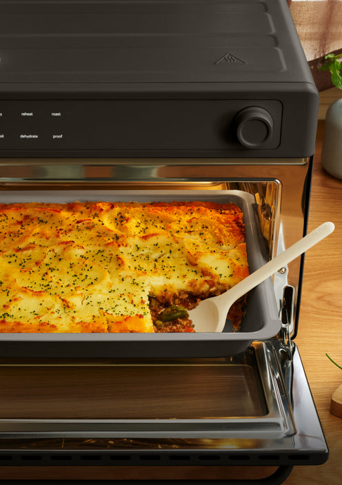 A shepherds pie in a baking dish inside a toaster oven, with a serving spoon scooping out a portion.