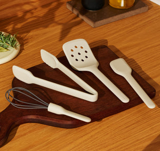 A set of light-colored kitchen utensils, including a whisk, tongs, a slotted turner, and a spatula, resting on a dark wood cutting board.