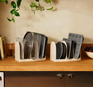 Two beige kitchen organizers on a wooden countertop holding various baking sheets, cooling racks, and muffin tins.