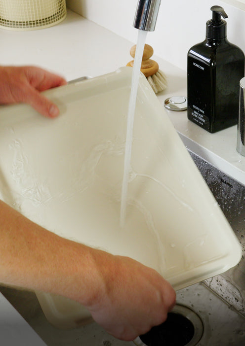 Hands rinsing a shallow, light-colored tray under a kitchen faucet.