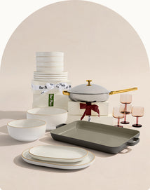 A set of white and gold-rimmed dishes, bowls, and glasses, with a lidded pan and a baking dish, arranged near stacked boxes tied with ribbon.