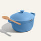 A Ceramic Nonstick Perfect Pot 6 L in blue, featuring two handles and a matching lid, sits on a light surface casting a shadow, with a wooden spoon resting on top.