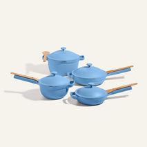 The Cookware Set features four blue nonstick pieces with wooden handles: two saucepans with lids, one stockpot with lid, and one sauté pan inspired by the Always Pan, all shown against a light background.