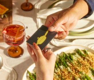 Two hands holding a card with a yellow duck illustration on a dining table with food and a drink.